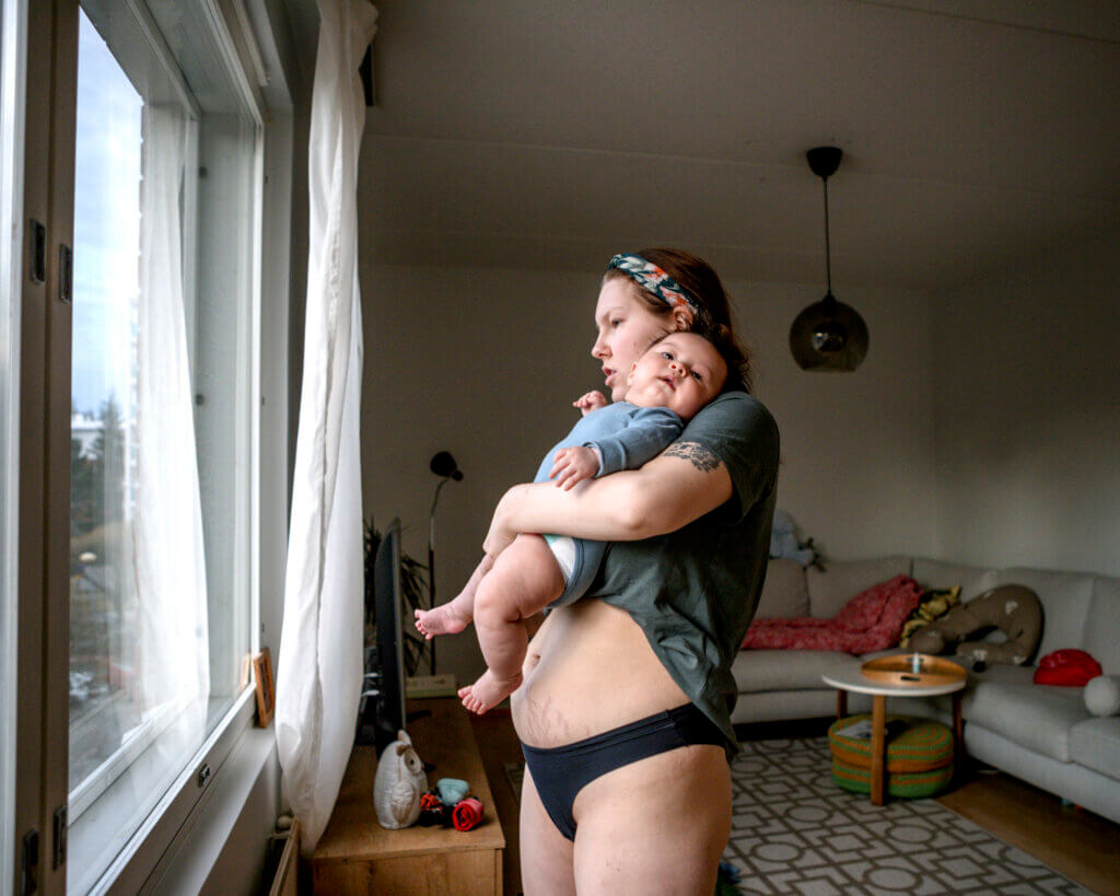 Nainen seisoo olohuoneessa katsoen ikkunasta ulos, kantaen vauvaa sylissään. Vauva katsoo kameraan.