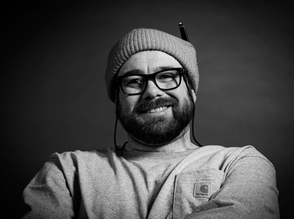 Hymyilevä henkilö pipo päässä ja silmälasit, harmaa paita. Mustavalkoinen kuva.