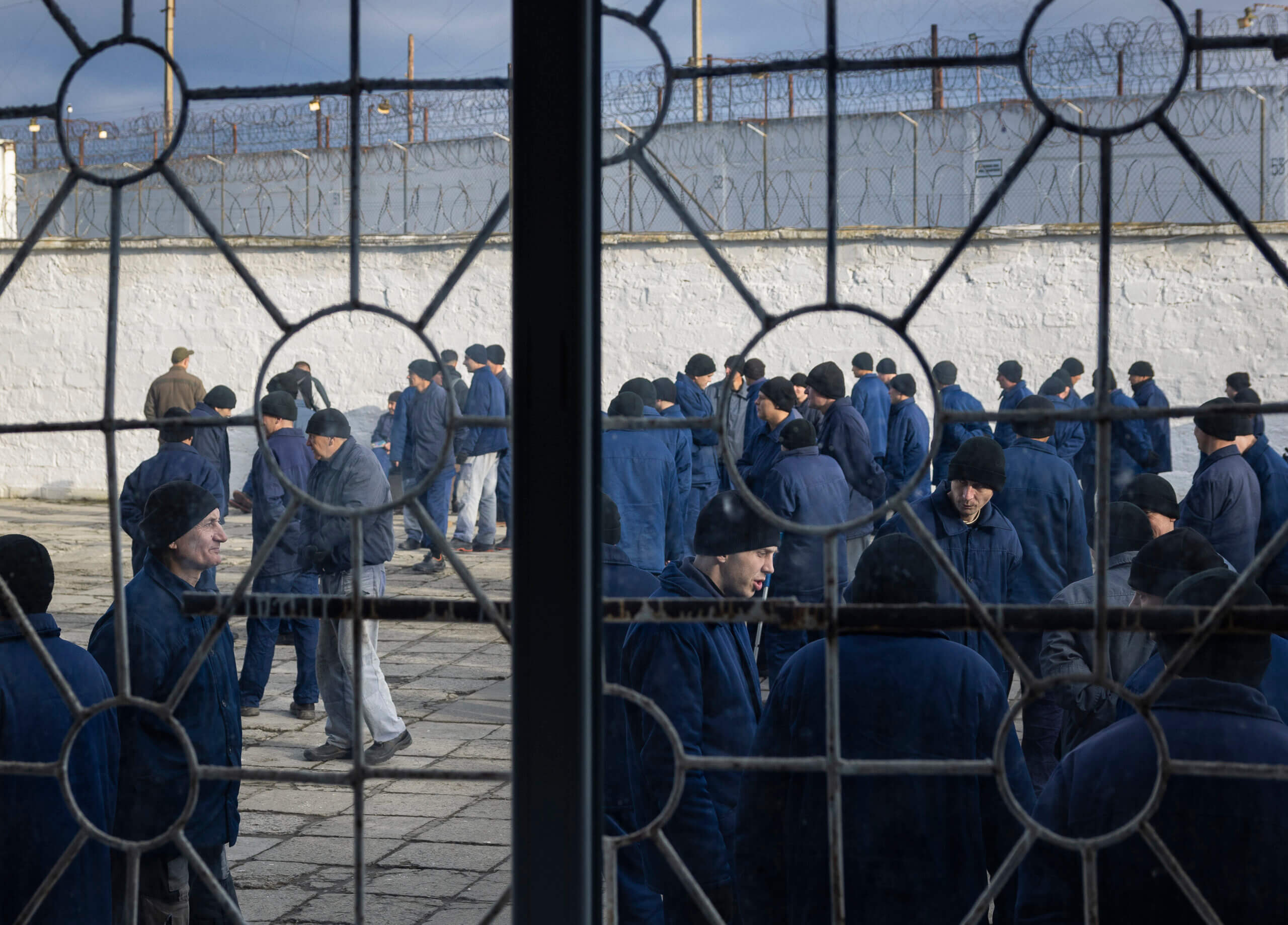 Vangit kävelevät pihalla piikkilanka-aidan ympäröimänä, kuvattuna ikkunan takaa.