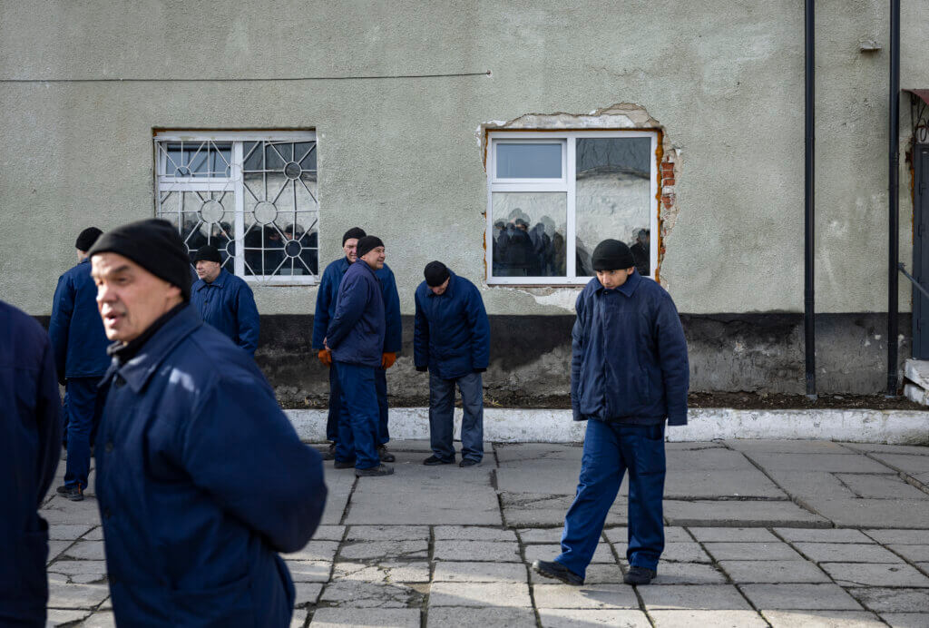 Joukko miehiä sinisissä vaatteissa seisoo ja kävelee betonialustalla harmaan rakennuksen edessä.